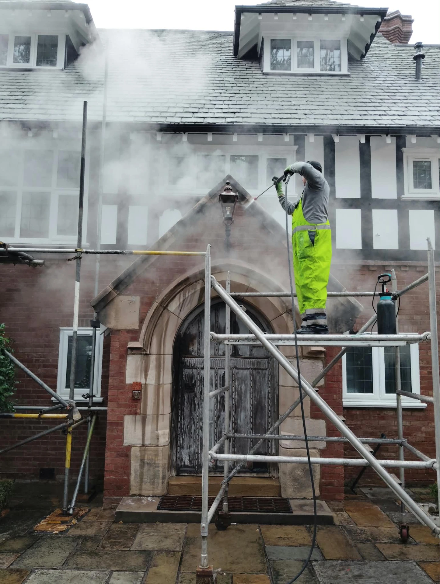 Operative steam cleaning the porch and render of a period Tudor-style house from scaffolding